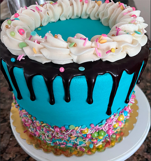 a round teal blue cake with a chocolate ganache drip and sprinkles surrounding the bottom of the cake