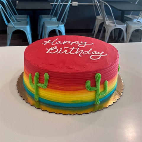 a round cake in red yellow and blue frosting with green cactus on the side