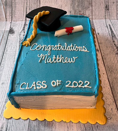 a graduation cake shaped like a book that had has a graduation cap and diploma on top. the cake also says Congratulation Matthew and Class of 2022