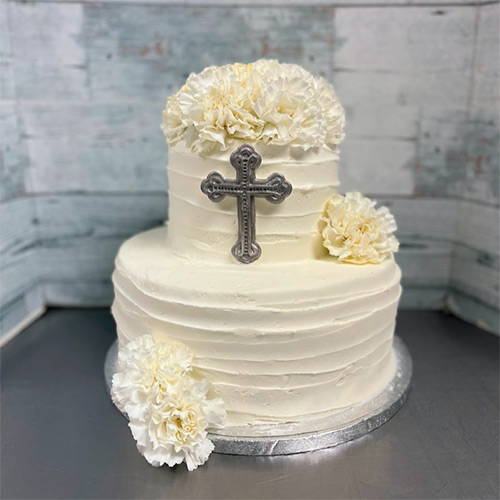 a two tiered white frosted cake with a silver cross on the side of the top tier