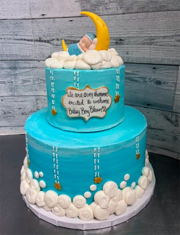 a two tiered blue cake with white clouds and a baby boy sleeping on a cresent moon on the top tier. on the side of the top tier displays the words We are over the moon excited to welcome Baby Boy number 2