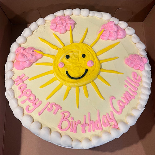 A round cake with a smiling yellow sun and pink clouds that says Happy 1st Birthday Camille