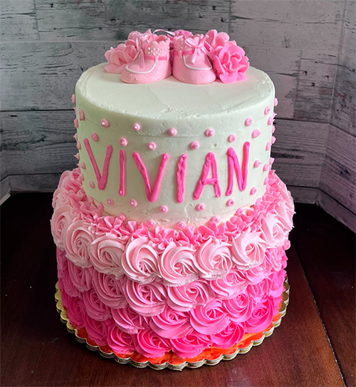 a two tiered cake with pink ombre flowers and pink booties on to with the name Vivian on the side of the top tier