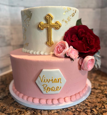 a two tiered pink, white and gold cake with a gold cross on the top tier and the name Vivian Rose written in gold on the bottom tier