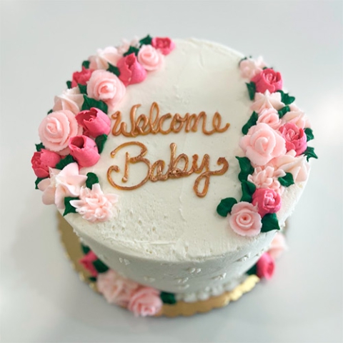 a round white cake thats displays the words Welcome Baby in gold and light and dark pink flowers around the top of the cake